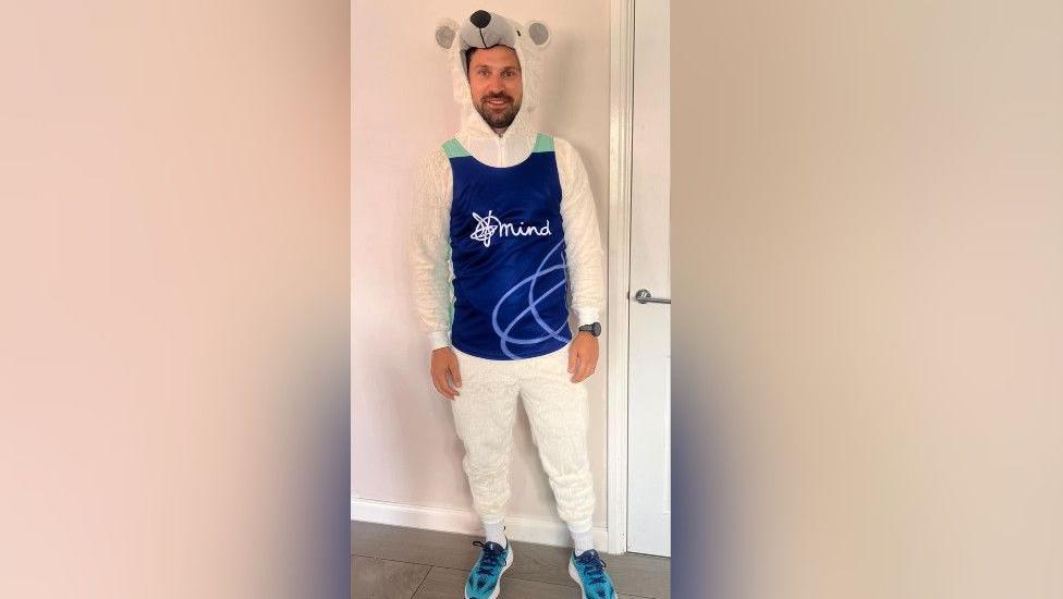 A full-length picture of Alex in the polar bear suit. It is white with a polar bear's head as the hood. He is wearing a dark blue vest that has a white Mind logo on it. He is wearing blue trainers.