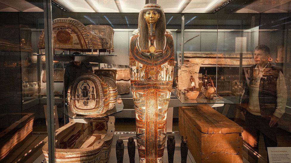 A highly-decorated Egyptian mummy case standing upright in a glass case with decorated sarcophagus to its right and a plain wooden box to its left. A man is looking through the glass at it and other ancient Egyptian artefacts can be seen behind it