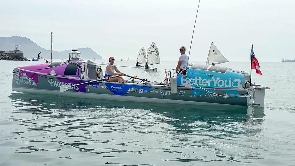 The two rowers sat in their boat which has two covered ends and a central open part. The hull is covered in sponsors names and a number of sailing ships are behind them.