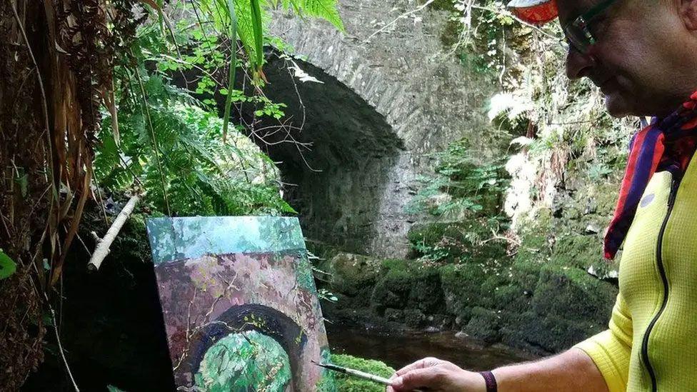 Timmy Mallett sits at an easel with a painting of a bridge on it in front of the bridge itself over a small river near Skyreburn