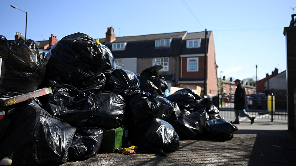 A big pile of black bin bags on a street on 2 April. A man in background walks past, with houses behind the bin bags