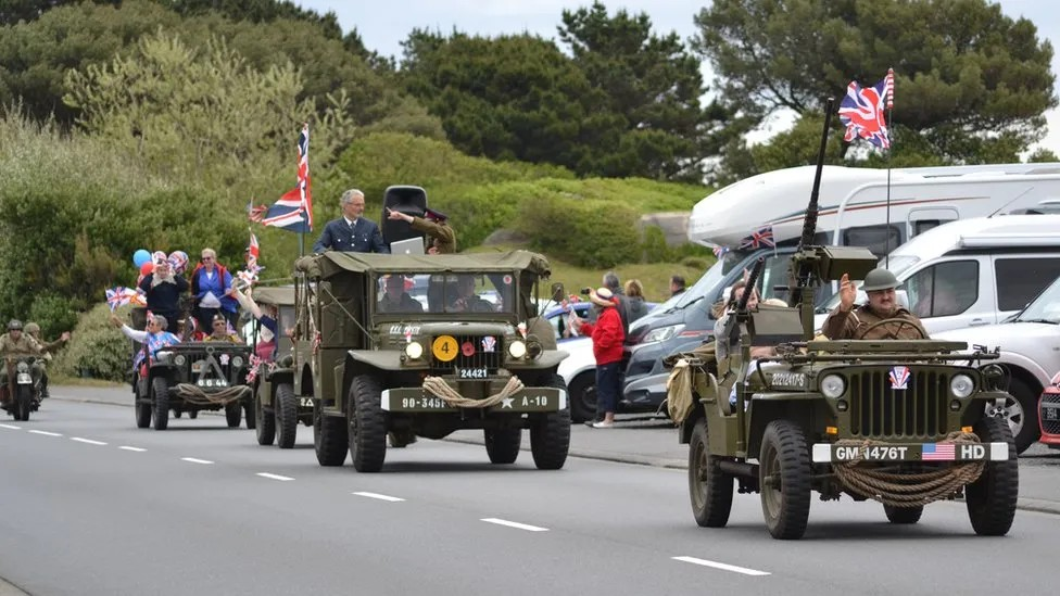 Liberation Day parade. Several World War 2 era vehicles decorated with flag, balloons and bunting in a procession along a main road in Guernsey. Drivers and passengers wear period costumes.