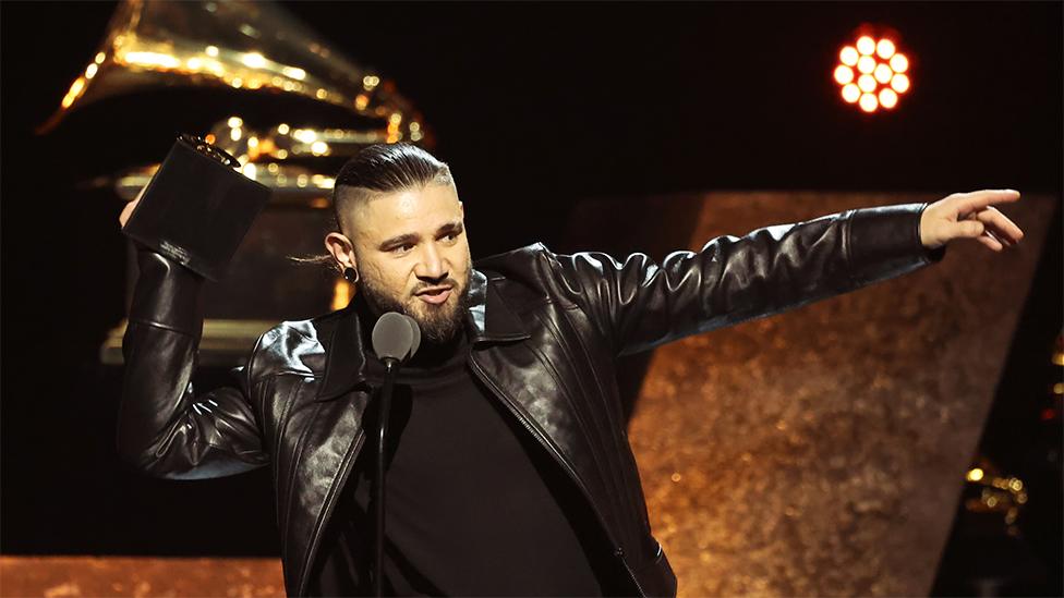 Skrillex wearing a black top and black leather jacket pointing to his left, while holding an award on stage at the Grammy awards.