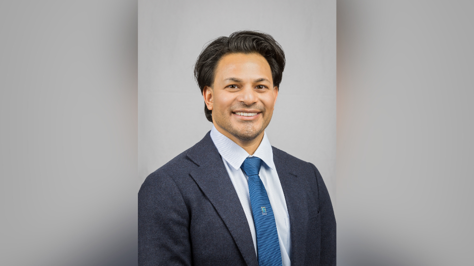 Photo of Dr Nuru Noor against a light blue background. He is smiling and wearing a dark blue jacket, light blue shirt and tie. 