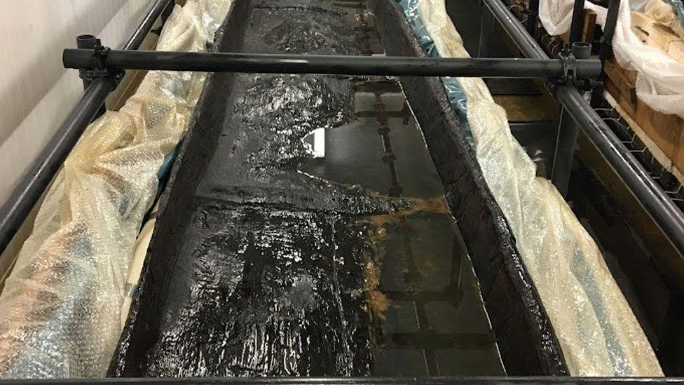 A blackened log boat in a bath of liquid in a conservation lab. The wood is mostly submerged and on either side its edges re protected with bubble wrap. 