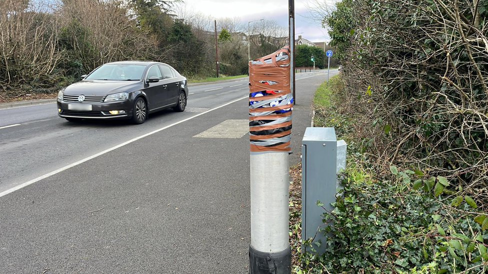 A stretch of the A4102 in Merthyr Tydfil. It appears the camera - in  layby -  has been broken off and the post has been taped up. Photo shows a black VW car driving past.