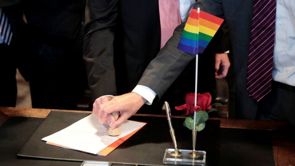 A same-sex couple stamp marriage documents on a desk with a rainbow pride flag on