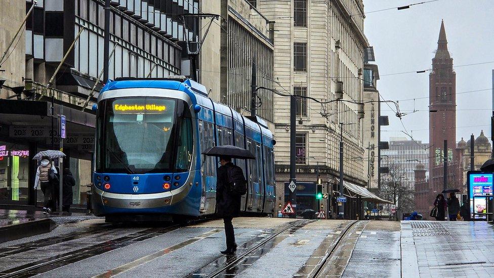 A tram outside a building in Birmingham city centre with a man holding an umbrella in the foreground and a clock tower amidst cloudy skies in the backgound