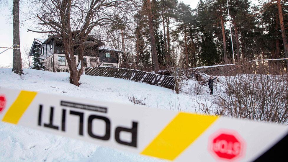 On a snowy hill, a home is seen surrounded by trees with a police tape across the foreground of the scene