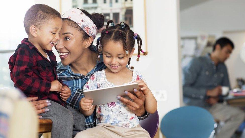 A woman plays with her two children at home with her partner in the background