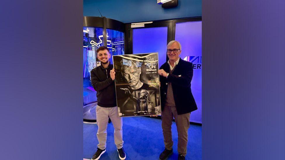 Artist Wilf Elliott and Avicii's father Klas Bergling holding up a black and white drawing of the late DJ. The artist is wearing a black zip-up jacket and cream trousers, and Mr Bergling is in a black jacket, white shirt, and beige jumper and trousers. They are in a room that has blue carpet and blue walls.