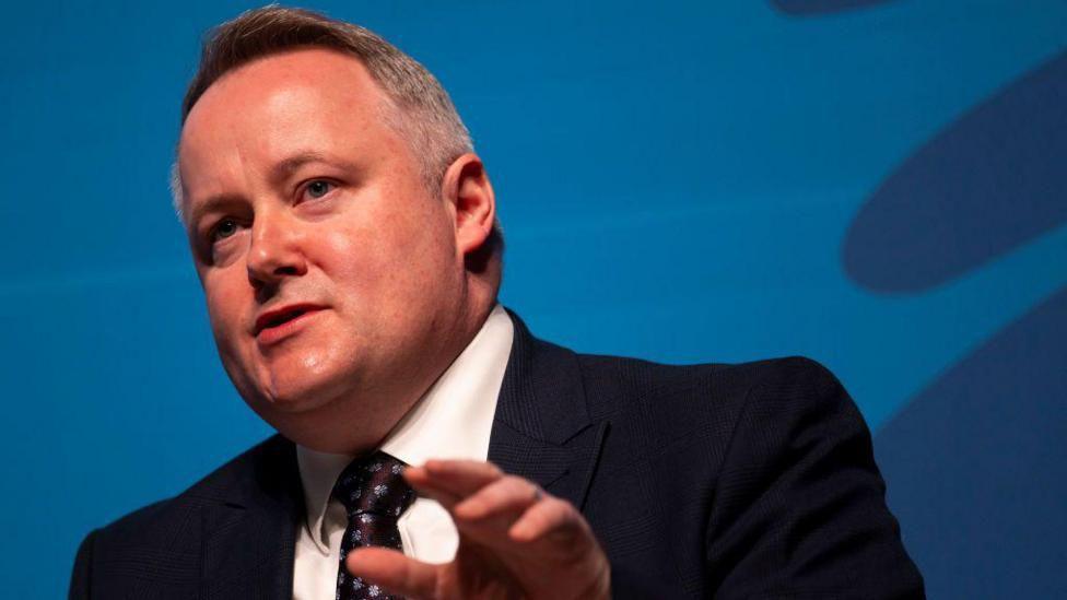 Darren Millar is stood with his head and shoulders in shot, gesticulating with his left hand. He is wearing a black suit, a tie and a white shirt, and is front of a blue background.