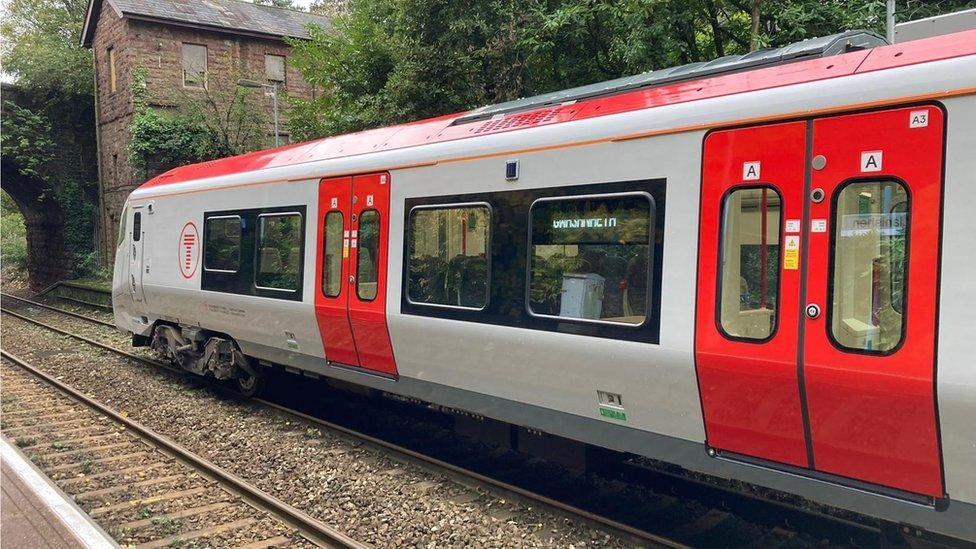 A Transport for Wales train in red and grey livery, on a rail track near a boarded up building