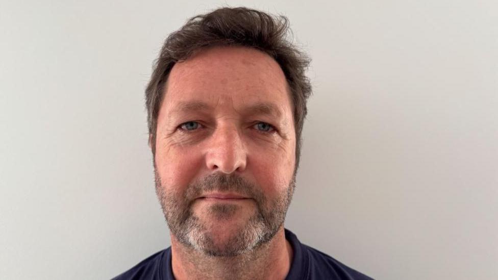 A man is smiling at the camera he has dark hair and is wearing a blue top 