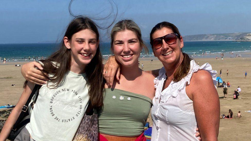 Ms Exton with her daughters Jess and Sophia stood arm in arm in front of a beach and the ocean.