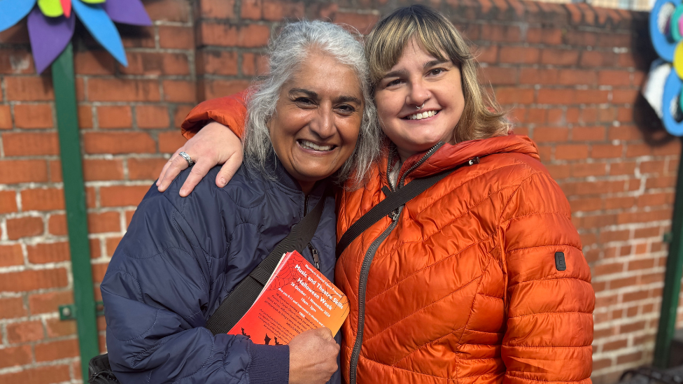 Bobbie in navy coat and Amy in bright orange coat smiling against red brick wall