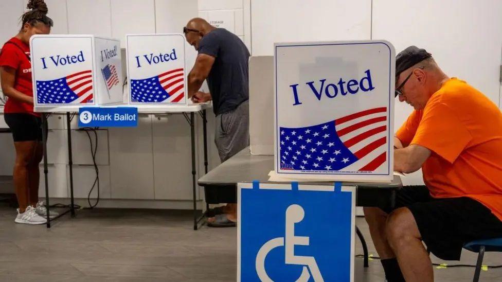 A voting center in the USA where a woman in a red T-shirt and dark trousers and a man with a blue T-shirt and grey trousers are standing at tables on which are ballot boxes with American flags and the words "I Voted" on the side. There is another table and ballot box to the right, which has a wheelchair symbol attached to it where a man in a blue cap, orange T-shirt and black shorts is sitting.