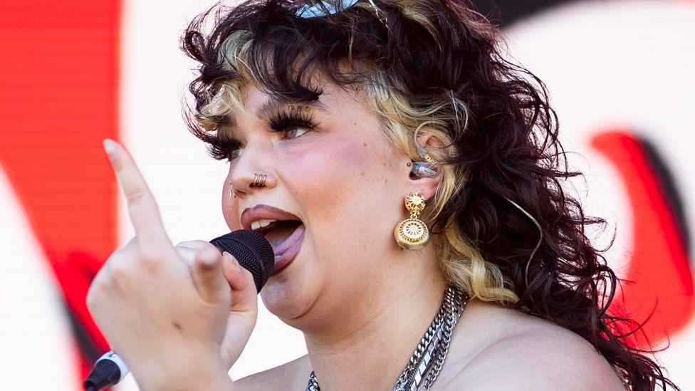 Lola Young performing on stage with a finger raised, in front of a red and white backdrop