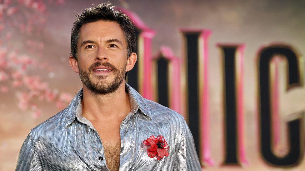 Joanthan Bailey in a shimmering silver shirt unbuttoned halfway down his chest in front of a giant Wicked sign at the film's premiere in Los Angeles on 9 November 2024