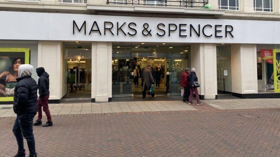 A view of the front of the M&S store in the centre of Ipswich. The white building has the name of the store above the entrance. Shoppers can be seen walking past.