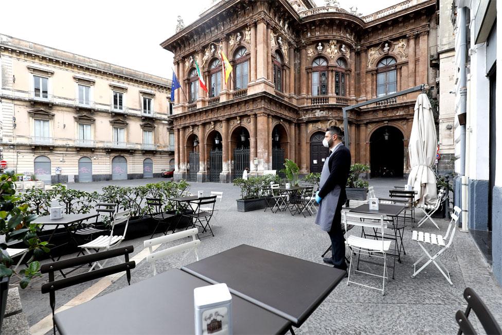 A waiter wearing a face mask stands outside a deserted restaurant