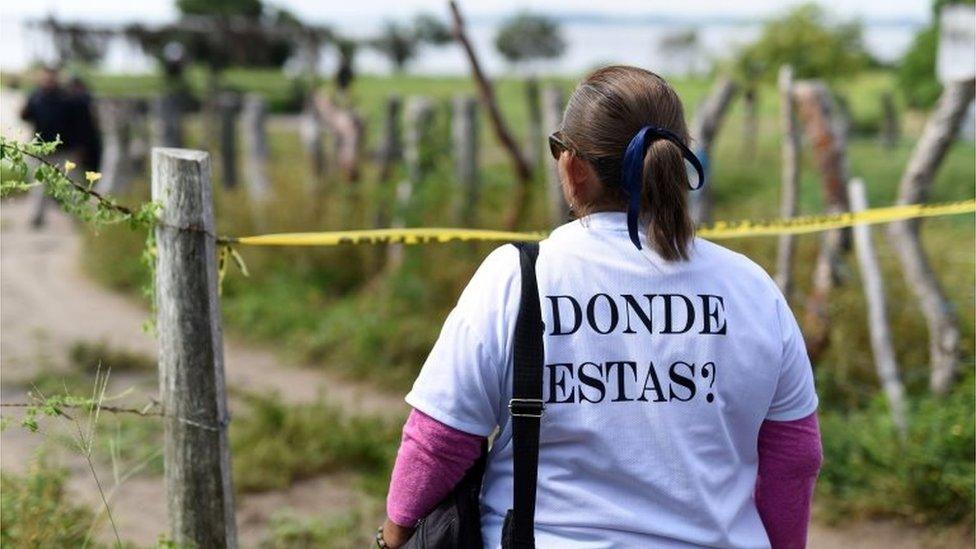 A woman wearing a T-shirt reading "Where are you?" looks at a site being searched by police