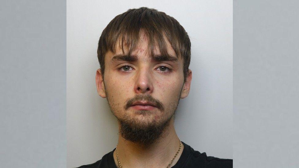 A police image of Cassius Adamson who is wearing a black top and silver chain. He is slim with some facial hair