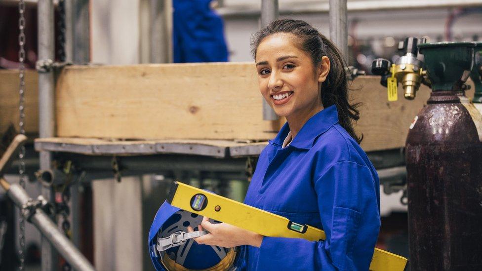 A female engineering student smiles as she holds a yellow spirit level and a blue helmet. She has long, straight, brown hair, worn in a ponytail, and is wearing bright blue overalls