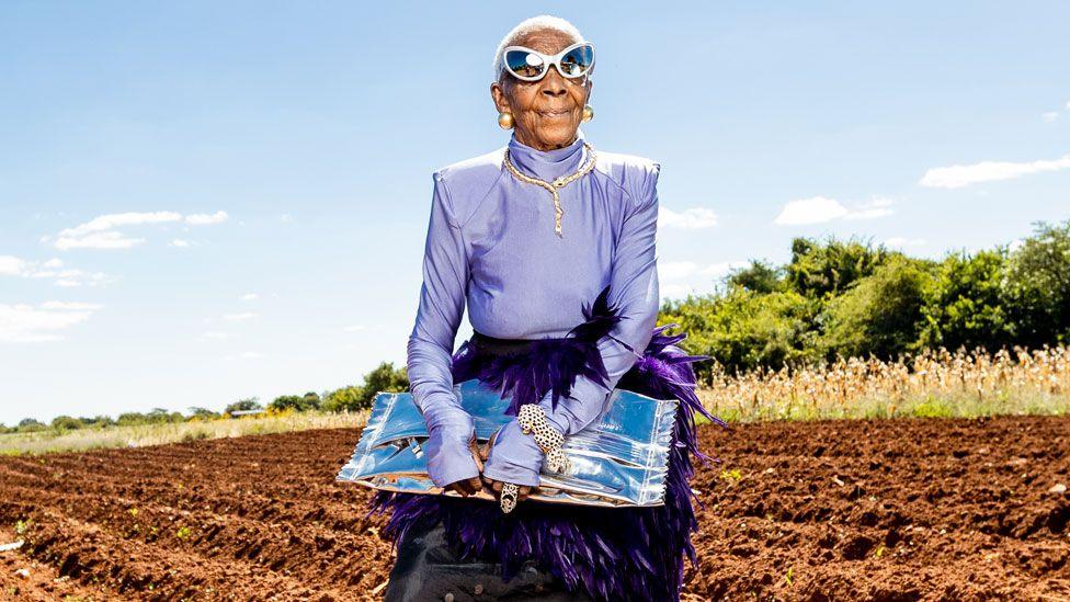 Margret Chola standing in a recently tilled field in a shiny lilac top with purple feathers around her waist and black skirt clutching a silver bag. She is wearing cat-eye mirrored sunglasses, a bejewelled gold necklace and bracelet in the shape of snakes and big gold ball earrings