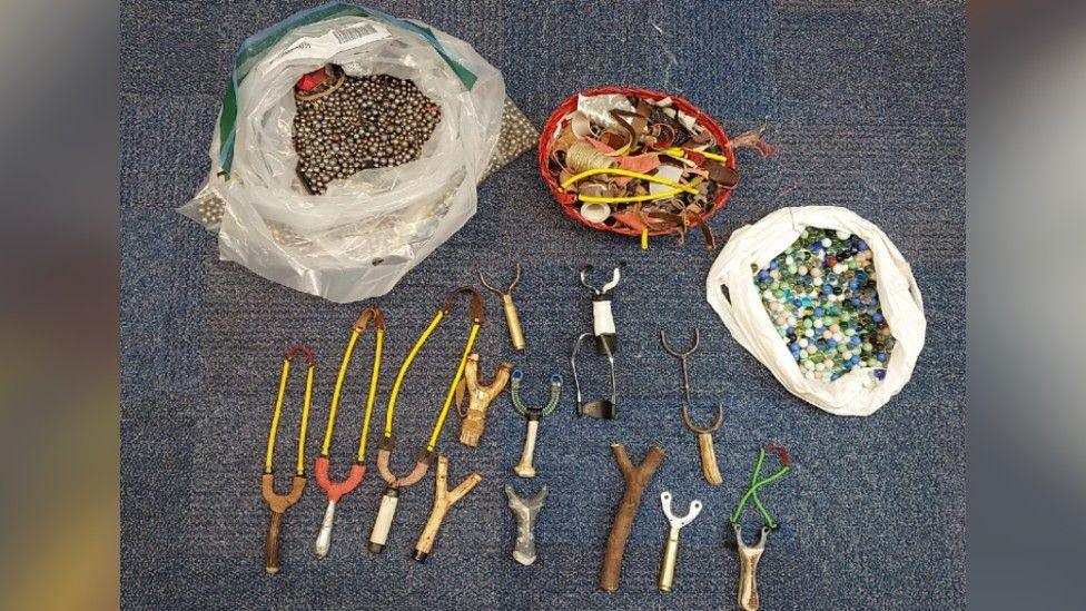 Fifteen catapults on a floor, next to bags of hundreds of silver-coloured ball bearings and marbles in bags. 