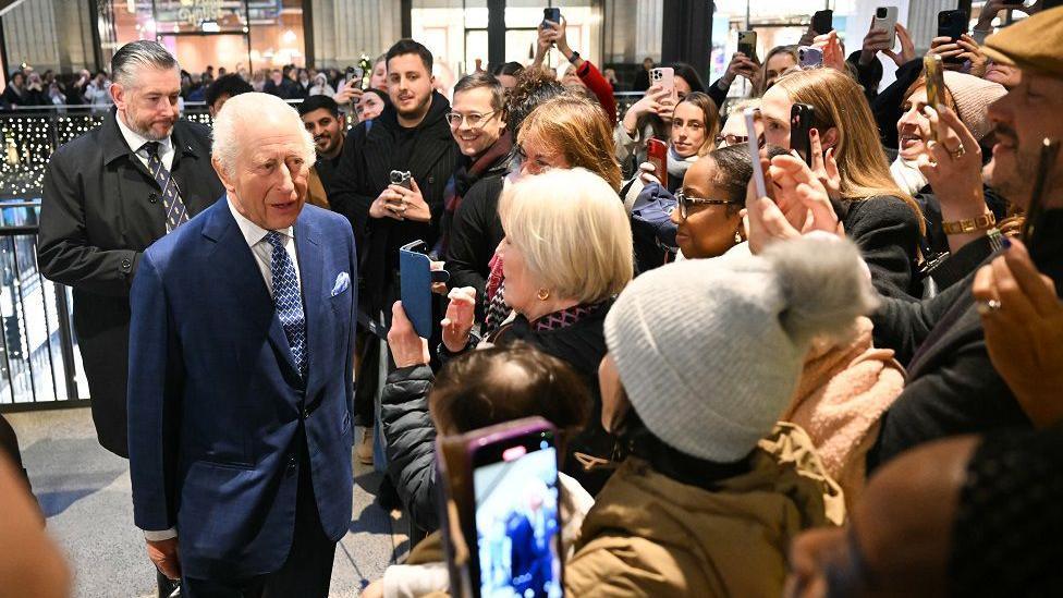 The King is surrounded by members of the public, many of whom have their phones out recording him