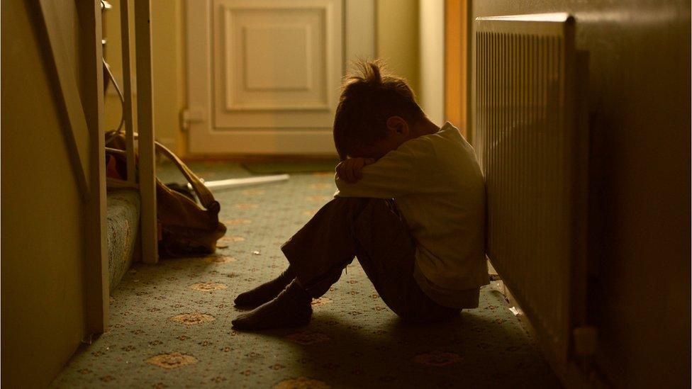 Boy sitting against a radiator in a hallway with his head down