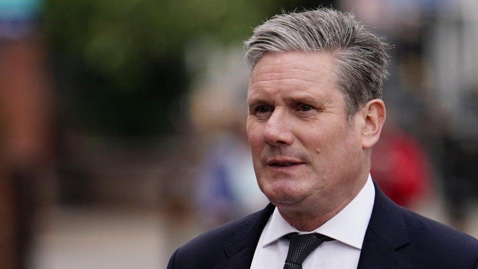 Sir Keir Starmer. He is stood outside, but the background is out of focus. He is wearing a dark blue suit and a black tie.