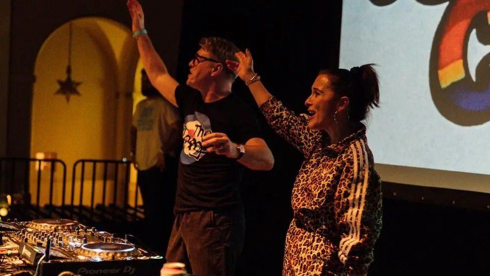 Johnny Owen and Vicky McClure are seen behind a DJ booth with their hands in the air.