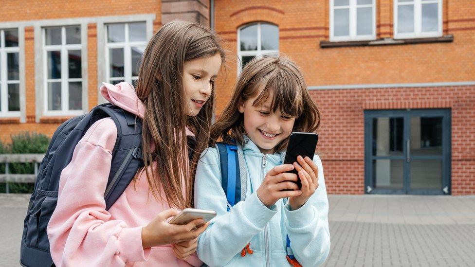 Two young girls use smart phones outside school.