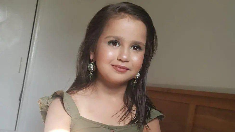 Sara Sharif with long, brown hair. She is wearing a green shirt, make up and earrings.