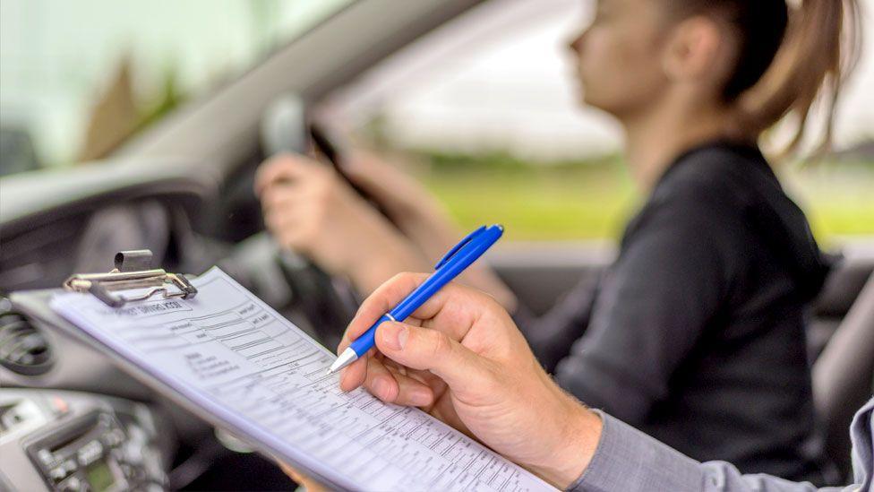 A driving instructor holds a pen to a clipboard in a car whilst a young woman takes her test in the driving seat