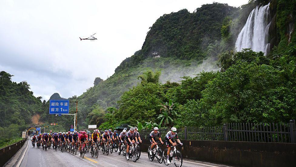 Cyclists race on a wet mountain road with lush greenery and a waterfall in the background. A helicopter hovers overhead, and a blue road sign with Chinese characters is visible above the cyclists.