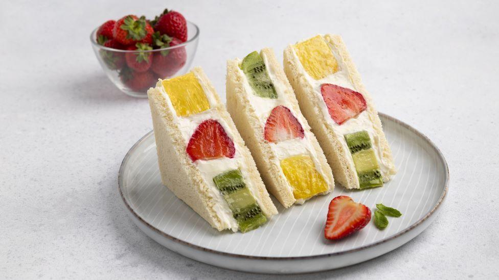 Three sandwiches in white bread on a plate. Each sandwich contains cream, a strawberry, a piece of kiwi and a piece of pineapple. In the background is a small bowl of strawberries