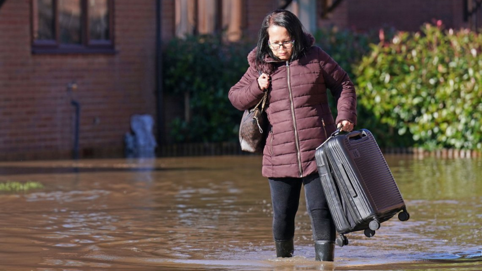 A woman with long black hair walks through flood water which is two-thirds up her Wellington boots. She wears a puffy magenta jacket with a handbag over her right shoulder and holds a suitcase in her left hand, raising it above the water.