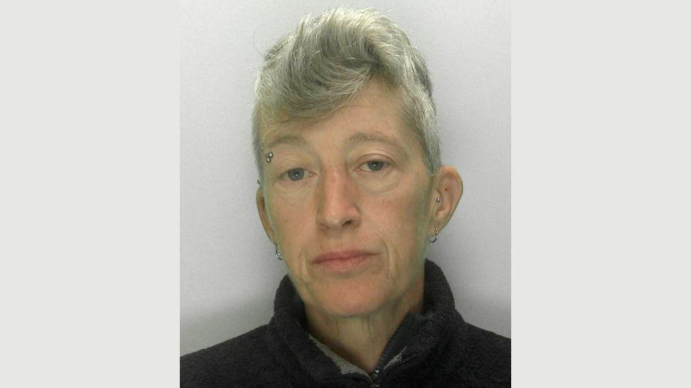 A police mugshot of Samantha Zala wearing a black pullover and standing against a grey background. She has short grey hair, an eyebrow piercing and two silver hoop earrings.