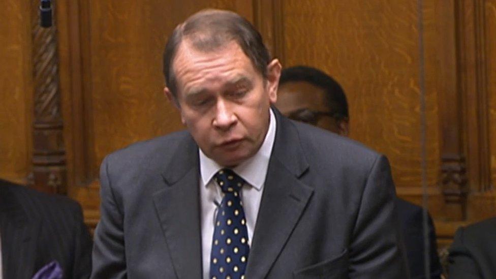 Man with short brown hair wearing a suit and tie stands up in the House of Commons