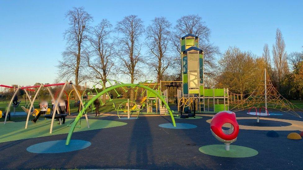 An image of a large green play area, with a large metal slide and netted bridge