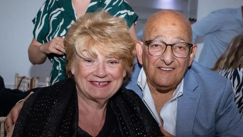 Elaine Marco has short hair and is wearing a black top and s smiling at the camera. Philip Marco is wearing glasses and a light blue suit jacket and white shirt and is also smiling at the camera.