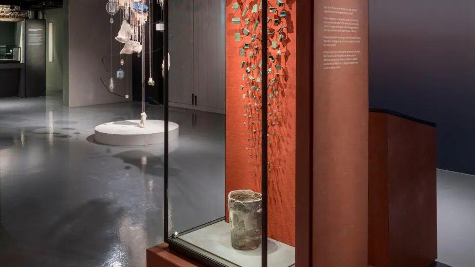 Items from an exhibition at Jersey Museum about the story of the island. The item at the front of the image is an old pot encased in a glass box with an orange display behind it. Pieces of pottery are on the wall of the display.