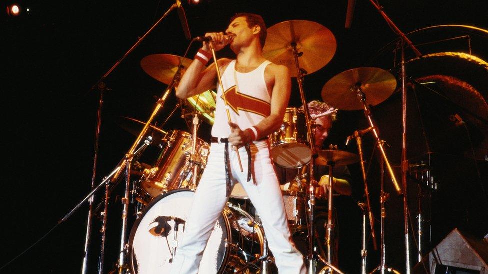 Freddie Mercury (1946-1991), singer with Queen, standing in front of a drum kit as he sings into a microphone on stage during a live concert performance by the band at the National Bowl in Milton Keynes, England