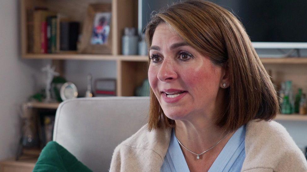 Kala Kilshaw looking away from the camera, with short brown hair, a silver necklace, light blue top with a cream cardigan over it. She is say on an armchair and there are shelves with books and trophies on in the background. 