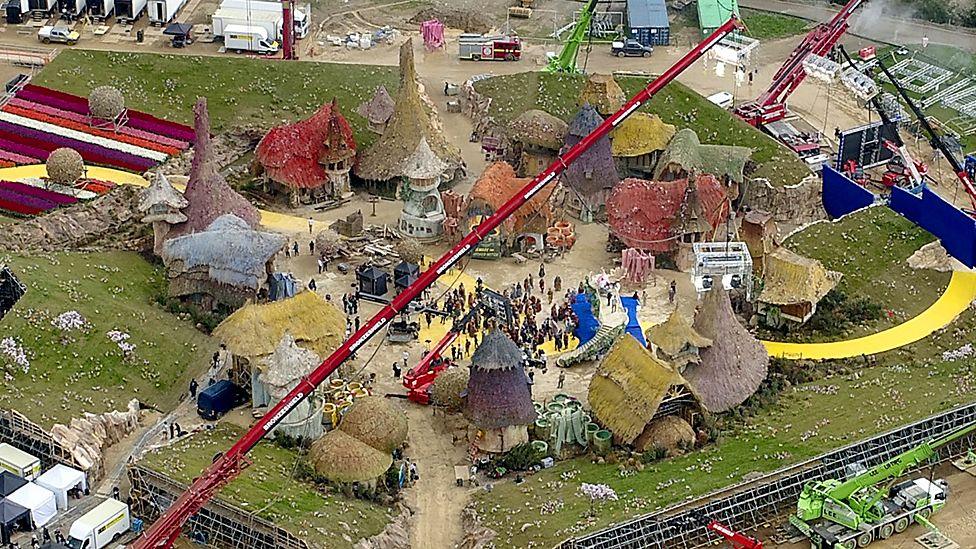 An aerial picture of the film set in Ivinghoe. It consists of a range of colourful, thatch-roofed homes all centred around a yellow brick road. The set is based in a field and there are various cars, trailers, cranes and other items associated with film production surrounding the set.