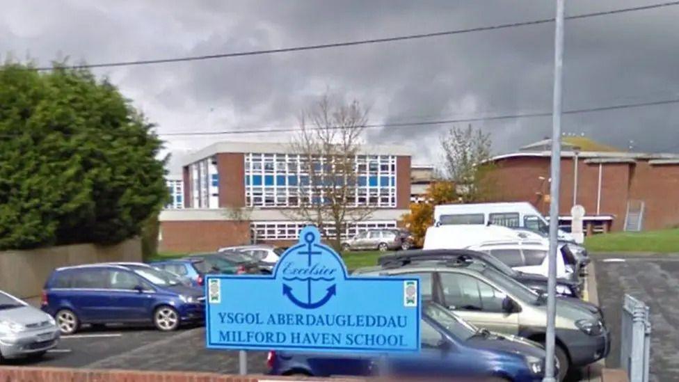 A photo of Megan Evan's school that she attended. There is a car park in the foreground with the school in the background. The school sign is also visible saying 'Milford Haven School'  and it's Welsh translation.