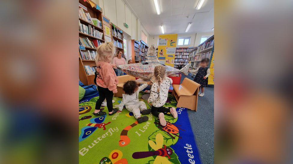 A multi-coloured model of a shark in the middle of a children's play mat in Woodston Library. There are four toddlers examining the shark, with a couple of adults in the background surrounded by bookshelves.